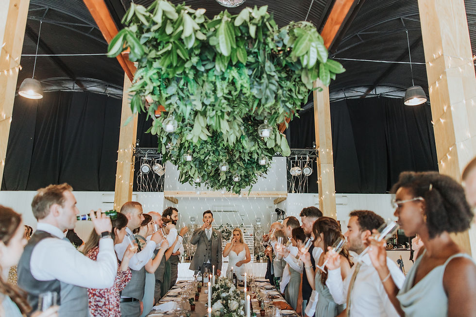 Wedding guests raising glasses at long banquet table with bride and groom, greenery ceiling installation above.