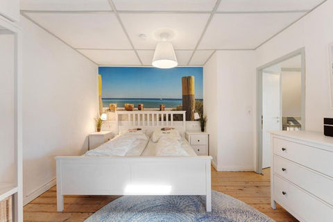 Schlafzimmer mit großem Doppelbett und weißer Kommode. Ein Teppich liegt auf dem Holzfußboden und die Wand hinter dem Bett ist mit einem Sandstrand und der Ostsee tapeziert.
