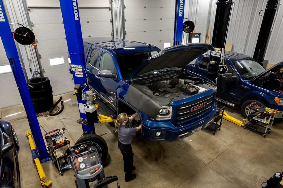 Female workingon passenger side of blue GMC sierra on lift. Blue ford f-150 on right side of image. Image includes 3 blue and yellow lifts. Grey car on left side of image.