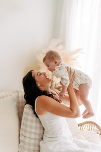 A joyful woman lifts a smiling baby in the air while sitting on a cozy sofa. Soft lighting and pampas grass create a warm, serene atmosphere.