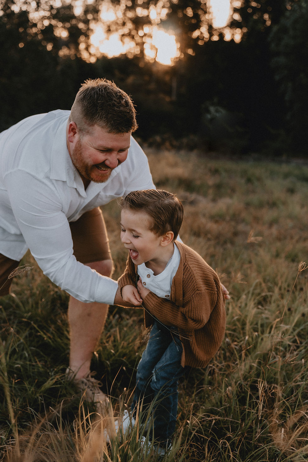 Father is smiling whilst tickling son in a white top and knitted brown jumper