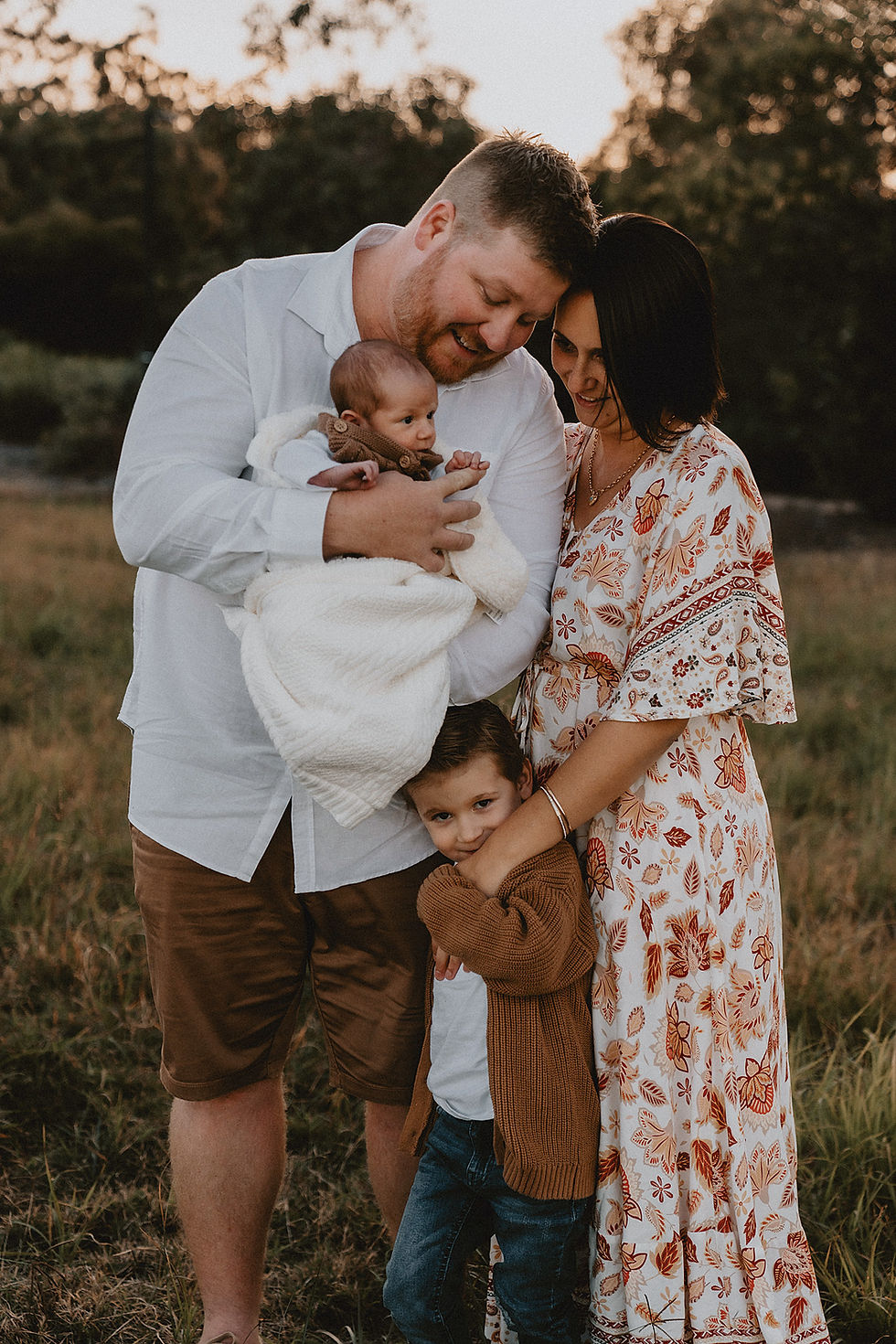 Father and Mother resting their heads together whilst holding newborn baby and an older child standing at their feet