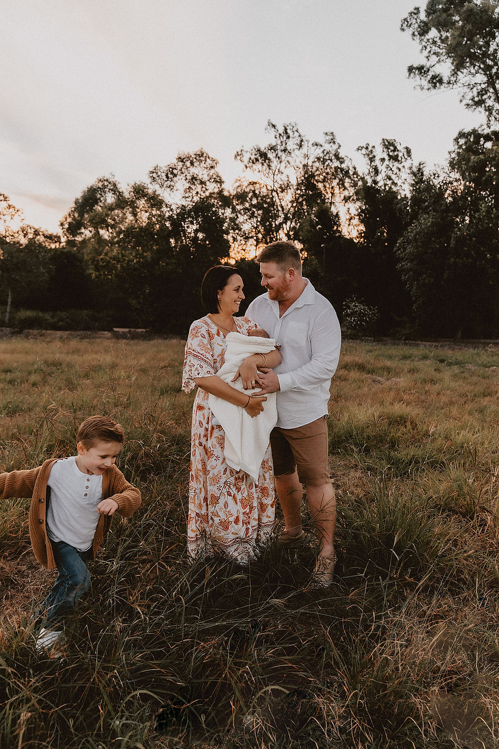 Mother holding baby while dad cuddles them both and little boy runs around them in field