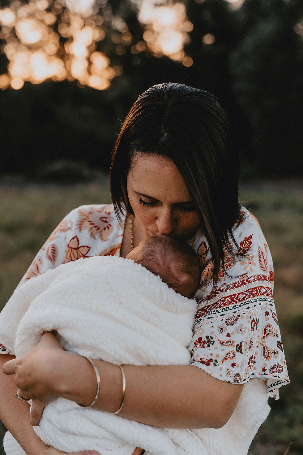 Mother holding newborn baby and kissing him on the head in a field with the sun setting behind them
