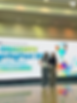 A wide shot of two colleagues at a medical conference; the background features a large green and blue banner that reads 'Longevity SpringFest 2026: Medicine Redefined.