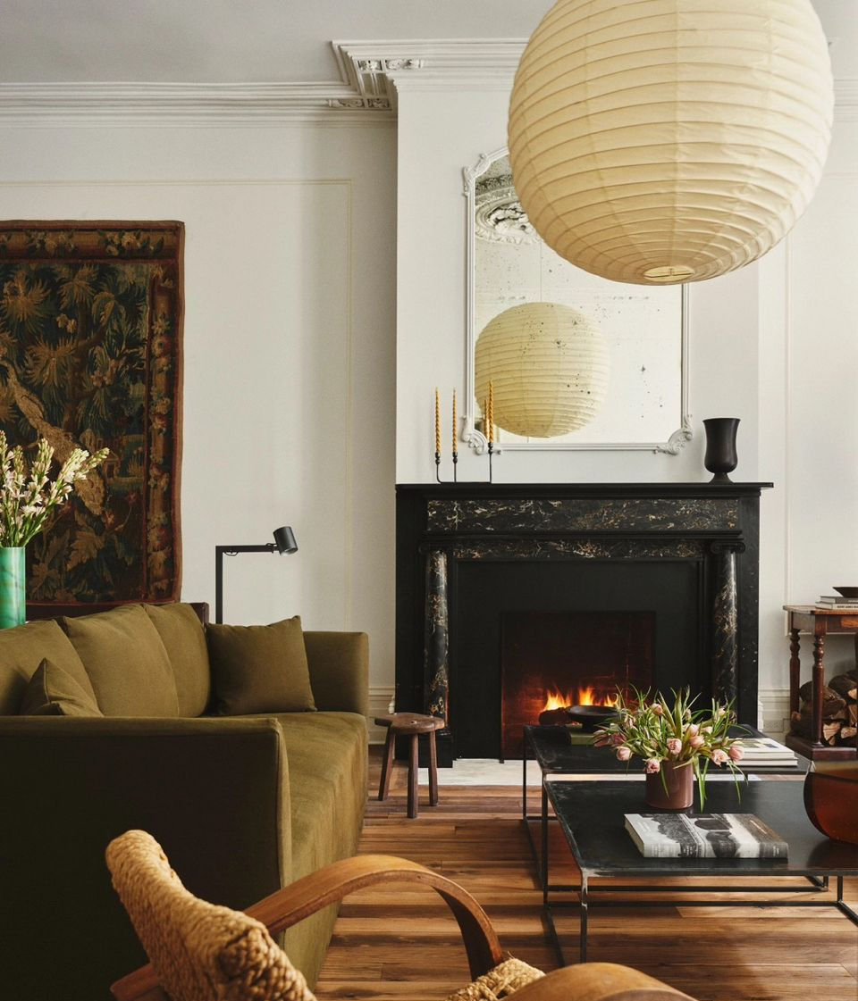 Salon chaleureux avec canapé vert, cheminée allumée, suspension en papier crème. Décor rustique avec tapisseries et fleurs. Ambiance cosy.