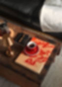 Tasse rouge à café sur livre beige à motifs rouges, table en bois, lampe dorée, canapé noir en cuire avec plaid blanc. Ambiance cosy et décontractée.