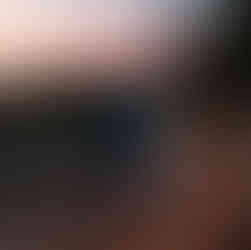 The elopement couple hikes along a dirt trail. She walks in front wearing her elopement dress and a matching white coat. He hikes behind wearing his suit and a stylish blue backpack. The sunset paints pink hues in the background.