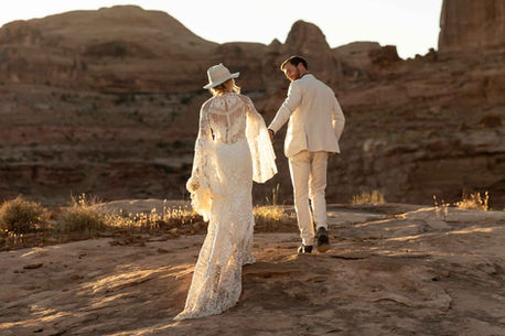 A couple eloping in Moab UT walks toward a red cliff at sunset
