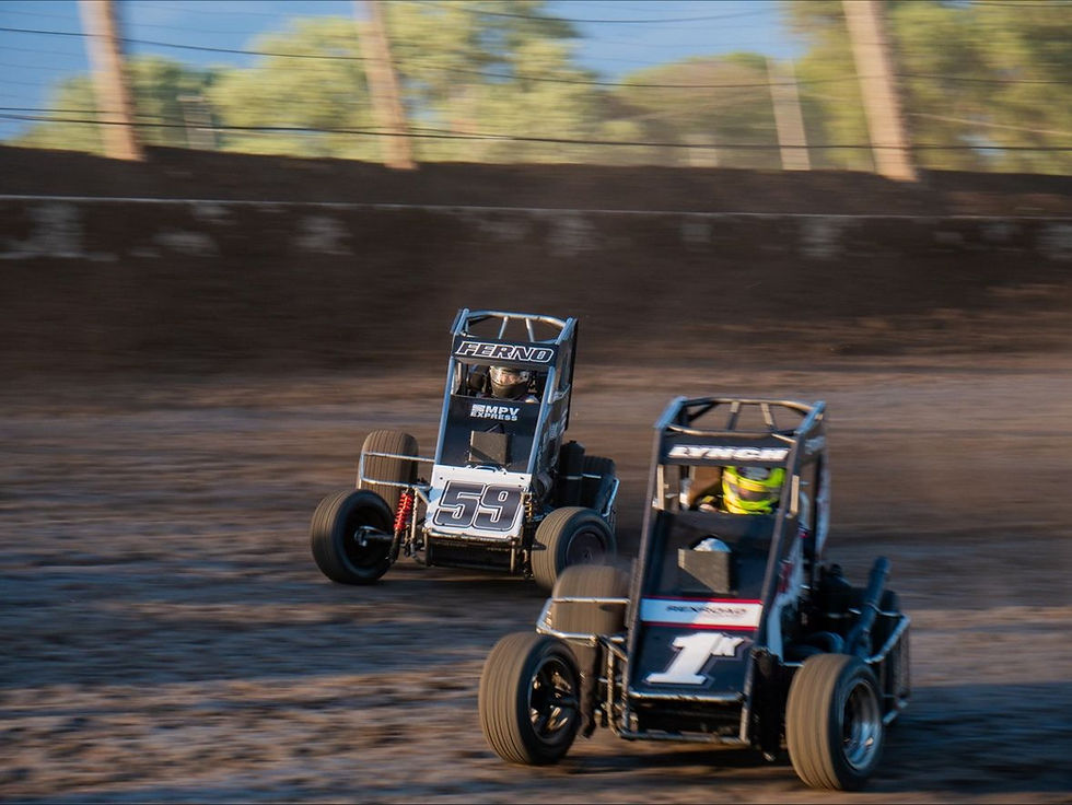Two sprint cars, labeled 59 and 1, race on a dirt track with blurred trees in the background. The scene conveys speed and competition.