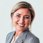 A woman with light brown hair, wearing a gray blazer, smiling in front of a light blueish-grey background.