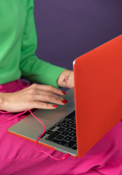 A person typing on a laptop with a red cover, dressed in a green sweater and a pink skirt, set against a purple background.