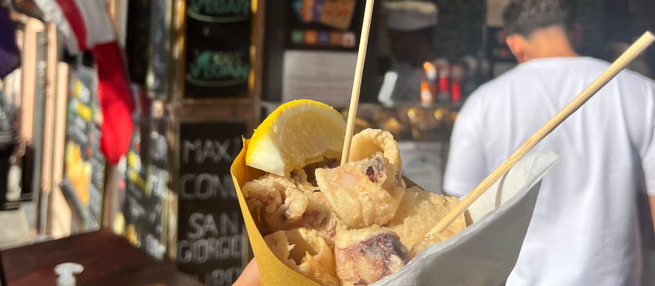 Hand holding fried food in a yellow cone with a lemon wedge, in front of a food stall. Flags hang above, with signs and text in the background.