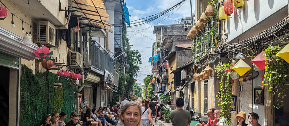 Bustling street scene with a smiling woman in the foreground. People sit along the stone path lined with vibrant lanterns and plants.