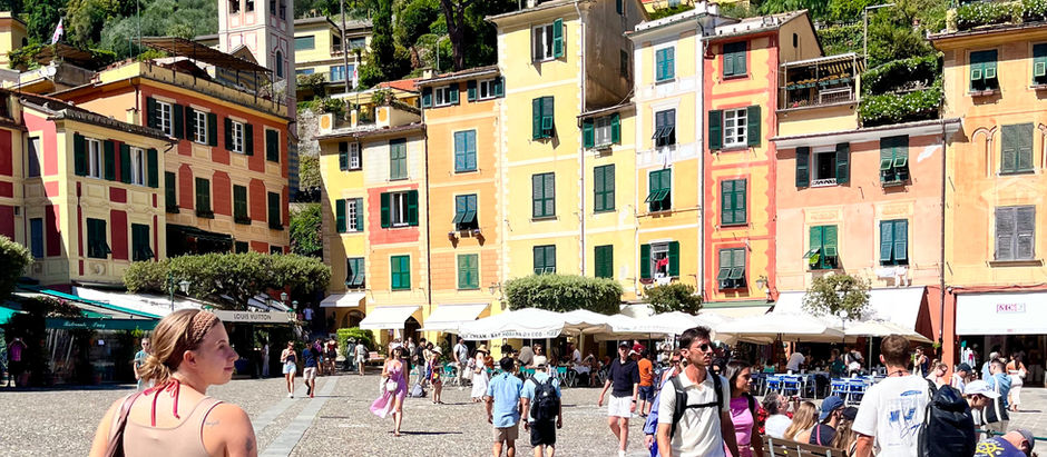 People walk on a lively cobblestone square with colorful buildings in a sunny coastal town. Green hills and a bell tower are in the background.