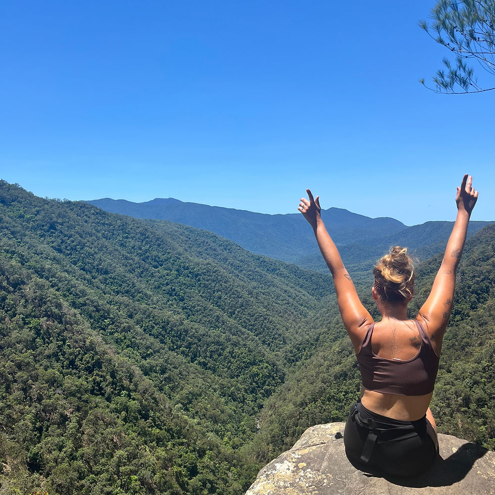 Woman sitting on a cliff with arms raised, overlooking a vast green valley and blue mountains under a clear blue sky. Feeling triumphant.