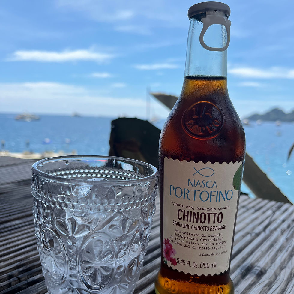 A bottle of Niasca Portofino Chinotto and an empty glass sit on a wooden table, overlooking a calm sea under a blue sky.