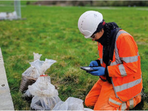 Ground investigation engineer in high-visibility clothing and a hard hat examining soil samples beside a site vehicle in a grassy field.