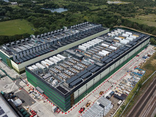 Aerial view of a large data centre facility under construction, featuring extensive rooftop cooling and power infrastructure, surrounded by service yards, railway lines and green countryside.
