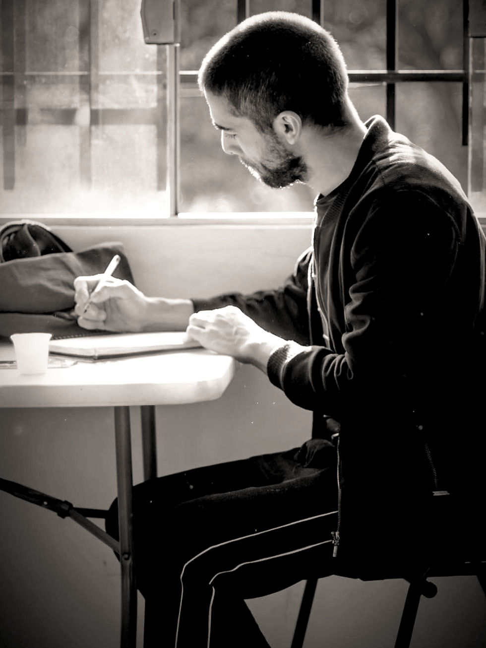 A imagem em preto e branco mostra um homem, “Thiago Ozelami, sentado a uma mesa registrando anotações para o mapeamento do cenário que aconteceria em breve, focado em escrever em uma folha de papel à sua frente. preparação e planejamento antes de um ensaio de teatro Ele veste uma jaqueta escura e parece pensativo antes do ensaio com os atores, com o corpo levemente apoiado na mesa. O ambiente é um espaço de ensaio, com uma janela ao fundo deixando entrar suave luz natural. para o homem, há um copo com água e outros papéis espalhados na superfície da mesa. A fotografia em preto e branco, feita por Giovanni Mattei, capta essa atmosfera concentrada e o processo criativo nos bastidores de um evento artístico.
