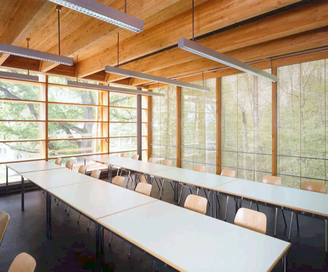 East facade with a vertical comprehensive shading element. Wooden slots are in between glass elements and block sunlight and avoid glare. The view into the surroundings is unobstructed.. Die Holzstäbchen sind die Glaszwischenscheibe eingelassen. Somit sind sie sogleich Sonnen- sowie Blendschutz. Die Durchsicht in den begrünten Aussenraum wird gewährt.