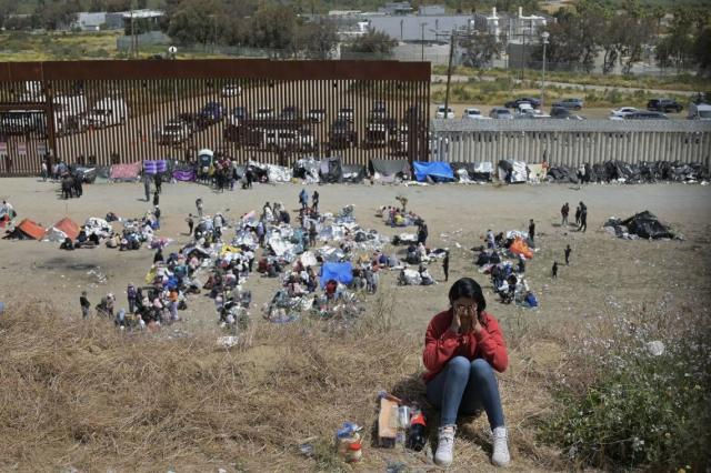 A distressed woman sits on a hillside near a border wall, with a large group of migrants and their belongings gathered below, highlighting the human impact of immigration policies.