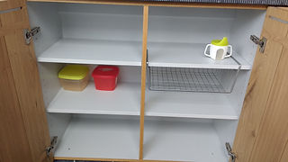 Open houten kast met witte planken, voedselcontainers, drinkbeker en draadmandje.