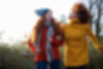 Two women, wearing knit sweaters, linked arm and arm, looking at each other and smiling.