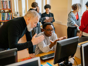 Teacher helping a student at a computer in a busy library setting, engaging in educational interaction.