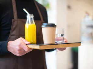 A barista serves juice and coffee on a wooden tray in a cozy café setting.