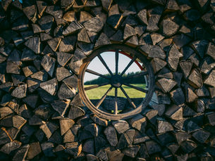 A vintage wagon wheel surrounded by neatly stacked firewood, revealing a lush green field.