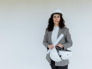 Engineer in formal attire holding blueprints and hard hats, standing against a white wall.