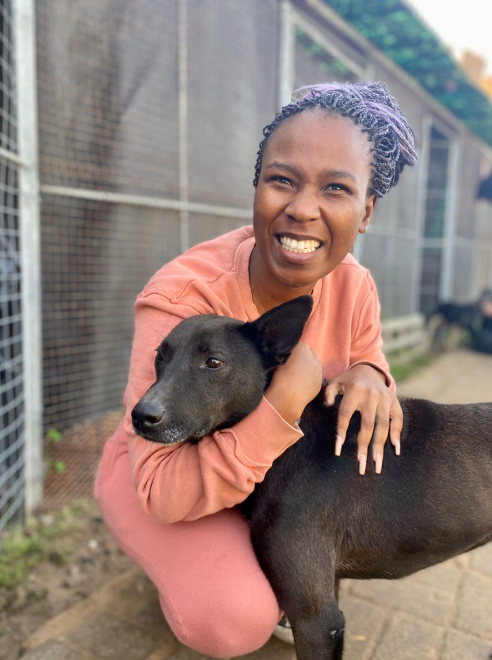 Smiling person in a pink outfit hugs a black dog outdoors, near a wire fence. The mood is joyful.