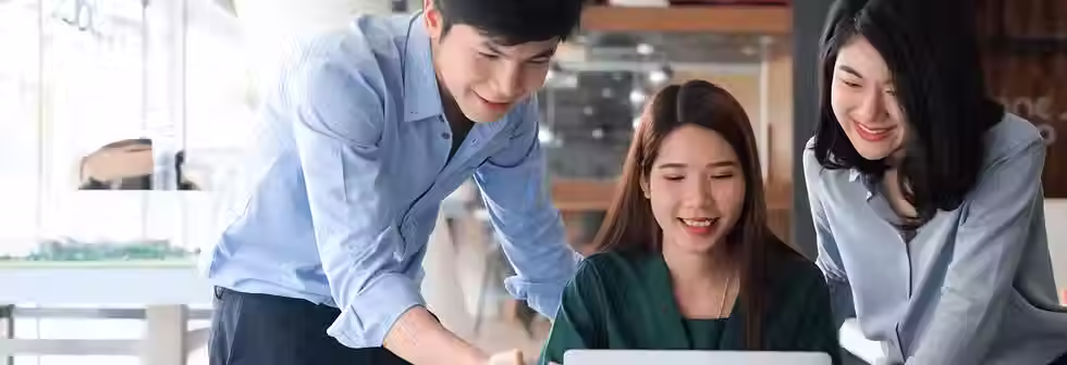 Three colleagues smiling and looking at a laptop in a bright office. Two women and a man, dressed in blue and green, collaborate cheerfully.