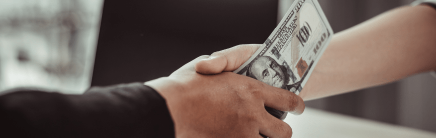 A person shakes hands with another while bribing them with cash, one of many examples of fraud