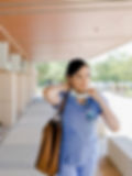 A woman in medical scrubs walks outside a building, carrying a tote bag and adjusting her face mask around her neck, reflecting the time management skills needed for busy healthcare professionals.