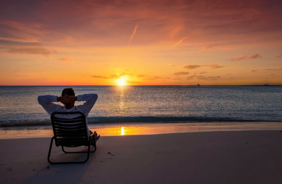 Person relaxes on a beach chair, watching a vibrant sunset over calm ocean. Sky is orange and pink, creating a peaceful mood.