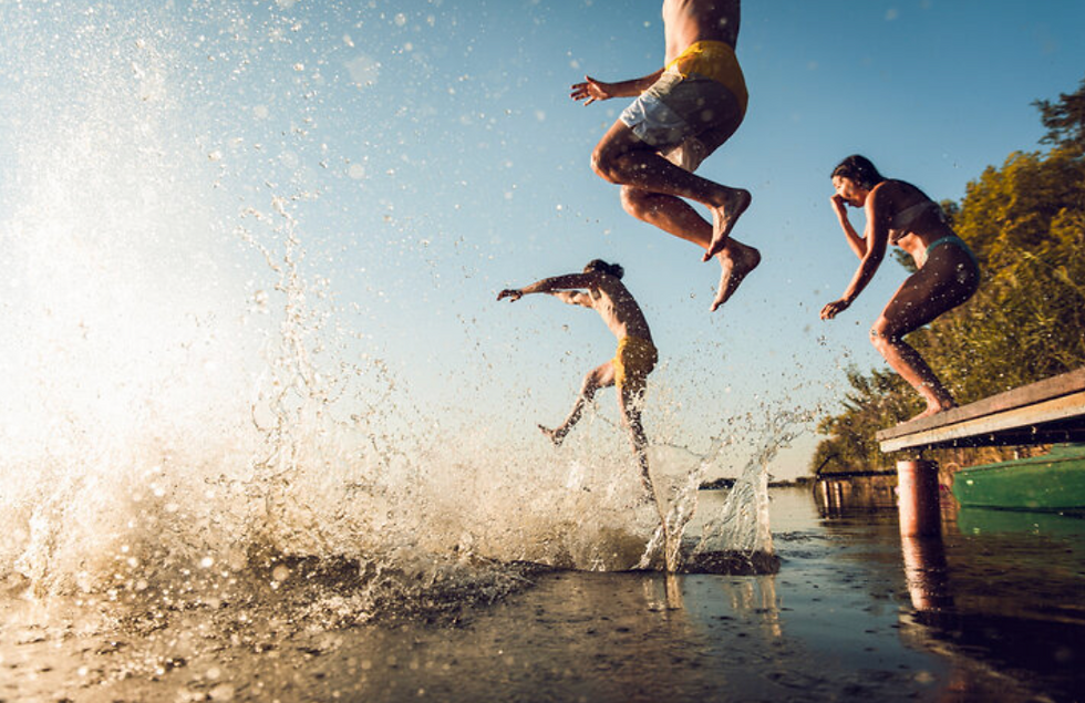 Three people jump off a dock into a lake, causing splashes. The setting sun creates a warm glow. Their expressions show excitement and joy.