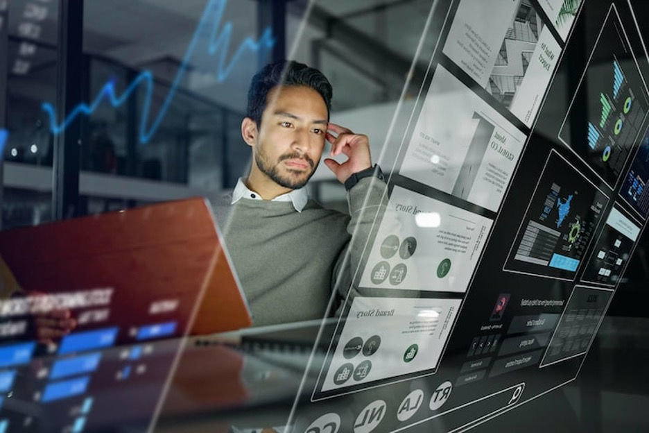 A man in a grey sweater sits at a desk in a modern office, looking thoughtfully at a large, angled holographic display. The transparent screen shows various UI elements, such as business presentations, infographics, and a search bar. In the background, blue neon line graphs float in the air, while the office's glass walls reflect the surrounding city lights at night.