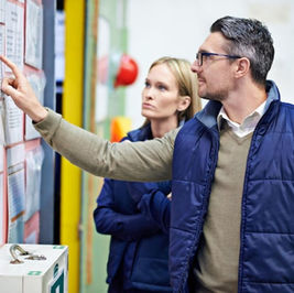 Man points at notices on a bulletin board while a woman watches in a school hallway, both in blue vests.