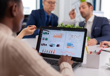 Person in office meeting points at a laptop with colorful charts while colleagues talk in the background.