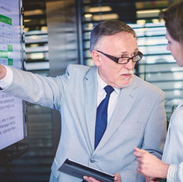 Two business professionals review a data dashboard on a large screen during a discussion about the modern lifecycle of Dynamics GP, with one presenter pointing to reports and analytics while holding a tablet.