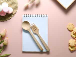 Flat lay of a notepad with a wooden spoon and fork on top, surrounded by tulips, pastel macarons, assorted cookies, and a pink background.