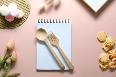 Flat lay of a notepad with a wooden spoon and fork on top, surrounded by tulips, pastel macarons, assorted cookies, and a pink background.
