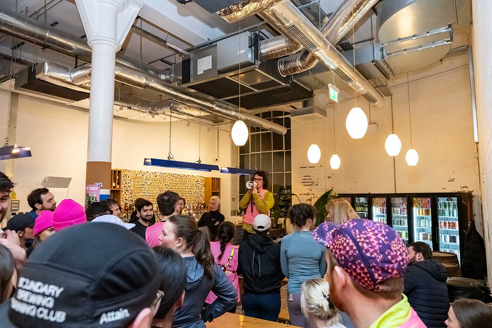 A person with a megaphone speaks to a crowd in a bar. People wear bright pink and black. Hanging lights and industrial decor set the mood.