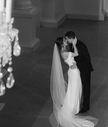 Black and white wedding photo of Sara and Steve sharing a romantic kiss in the Blue Room at the Henry Ford Museum. The bride wears a sleek white gown with a flowing veil, and the groom is in a classic black suit. A crystal chandelier in the foreground adds elegance to the historic venue. Wedding picture captured by Bearded Wolf Productions.