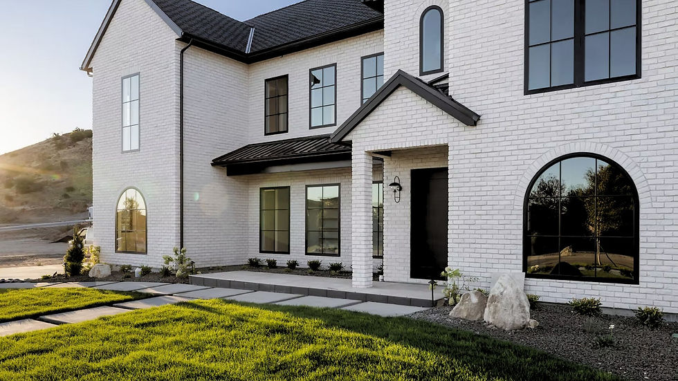 Modern white brick house with black windows and roof, green lawn in front. Sunlight casts a warm glow, hill and trees in background.