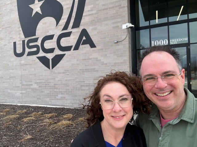 A smiling man and woman pose outside a building with "USCCA" on the wall. They're in front of a brick exterior and a glass door.