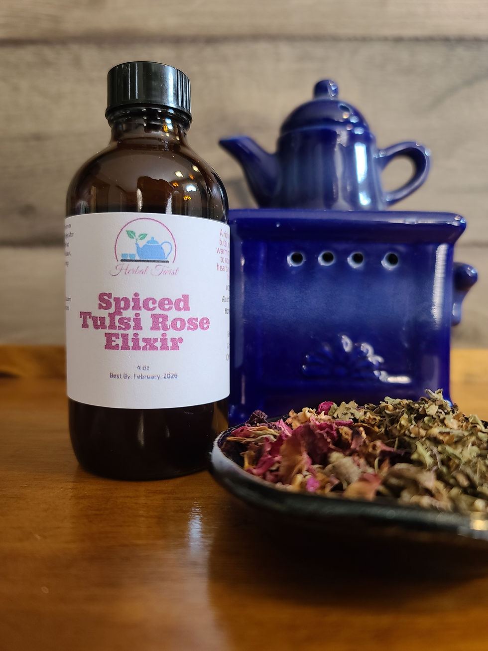 Brown bottle labeled "Spiced Tulsi Rose Elixir" on a wooden table, beside blue teapot and plate of dried herbs, evoking a natural, calming vibe.