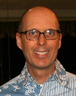 Alan Michael Smith. He is a light-skinned middle-aged man with a shaved head. He is wearing glasses. He is wearing a blue and white patterned shirt with stripes on one side and floral on the other.